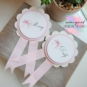 Puede incluir: Dos insignias de purpurina rosa y blanca con cintas. Una insignia dice "Mommy" y la otra dice "Daddy".