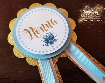 May include: A blue and gold rosette with a white circle in the centre. The circle has white polka dots and the word "Momma" written in gold glitter. The rosette has blue and white ribbons attached.