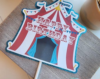 May include: A circus-themed birthday cake topper. The topper features a red and white striped tent design with the words "HAPPY BIRTHDAY" in white. It has a blue glitter border and is attached to a white stick.