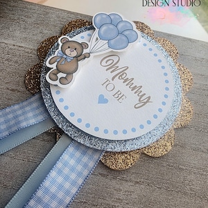以下が含まれることがあります： 青い風船を持ったテディベアのベビーシャワーロゼット。ロゼットは、金色のグリッターのスカラップエッジ、青色のグリッターの層、そして「Mommy to Be」のテキストが入った白い中心部があります。青と白のギンガムリボン。