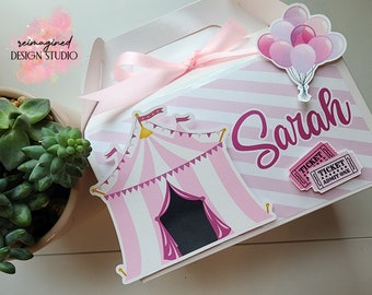 Op de afbeelding: Roze en wit gestreepte cadeaubox met een roze circustent, ballonnen en kaartjes. De doos is vastgebonden met een roze lint en heeft de tekst "Sarah" op de voorkant.