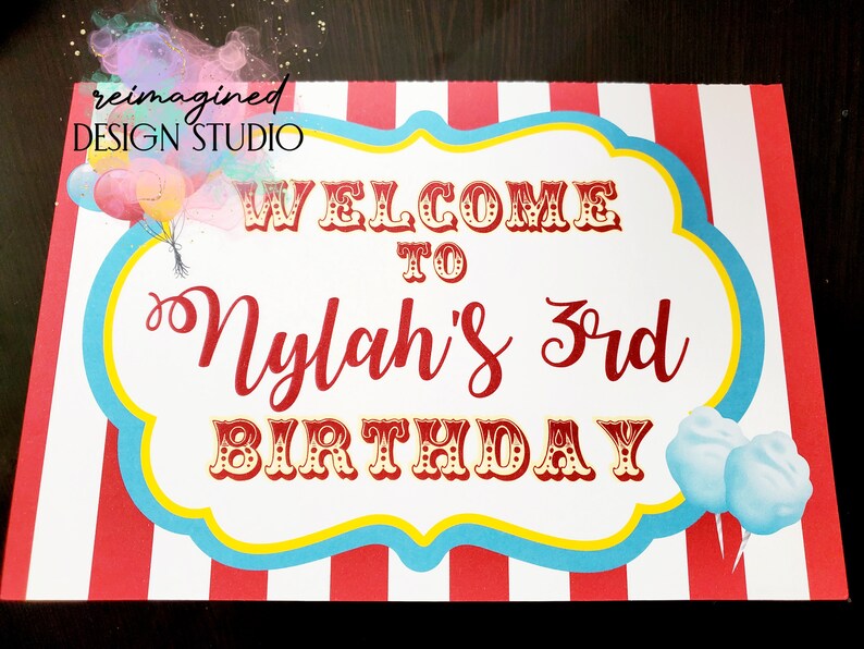 Peut inclure: Une pancarte blanche avec des rayures rouges et bleues autour du bord. La pancarte dit "Bienvenue &agrave; Nylah's 3rd Birthday" en lettres rouges et dor&eacute;es. Il y a des ballons color&eacute;s et de la barbe &agrave; papa sur la pancarte.