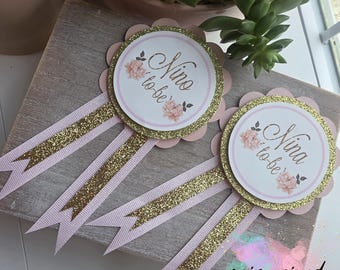 May include: Three pink and gold gender reveal party favors with gold glitter ribbon. The favors have a pink and white circle with the text "Nino to be" and "Nina to be" in gold lettering.