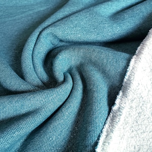 May include: Close-up of teal fabric with a textured weave, showing folds and a soft, light-colored edge. The fabric has a denim-like appearance, with a subtle sheen. The material is likely used for clothing or home decor.