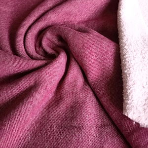 Pode incluir: Close-up de um redemoinho de tecido bordô com uma aparência de malha texturizada. Uma seção de tecido felpudo branco e fofo é visível no lado direito. O tecido parece macio e flexível, adequado para roupas ou têxteis domésticos.