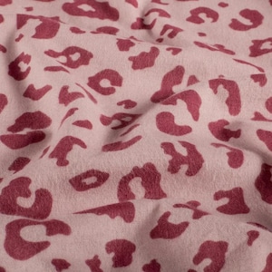 Può includere: Primo piano di un tessuto con sfondo beige rosato e motivo ripetuto di macchie leopardate color amaranto. Il tessuto è morbido e ha una consistenza leggermente stropicciata, suggerendo che si tratta di un materiale leggero.