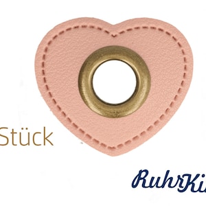 Könnte beinhalten: Zwei herzförmige Etiketten aus rosa Leder mit einem goldenen Metallösen in der Mitte. Der Text "2 Stück" ist unter den Etiketten gedruckt.