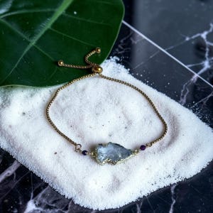 May include: A gold-toned adjustable bracelet featuring a light blue, irregularly shaped stone pendant. The bracelet has a delicate chain and is accented with small, dark purple beads. The jewelry rests on a bed of white sand.