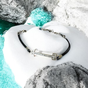 Puede incluir: Una pulsera de flecha plateada con cristales transparentes sobre un cordón negro. El diseño de la flecha es el punto focal de la pulsera, que se muestra sobre una superficie blanca con un fondo de agua turquesa.