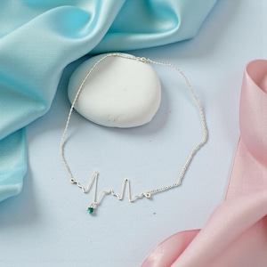 May include: A silver necklace in the shape of a heartbeat, with a small green gemstone pendant. The necklace is displayed on a light blue surface with blue and pink silk fabric in the background. The necklace is delicate and elegant.