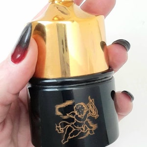 Puede incluir: Botella de perfume dorada y negra con un diseño de querubín. La botella tiene una tapa dorada y una base negra con un querubín dorado que sostiene una lira.