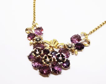 May include: A gold chain necklace with a purple and gold floral design. The necklace features a cluster of purple rhinestones and gold bows.