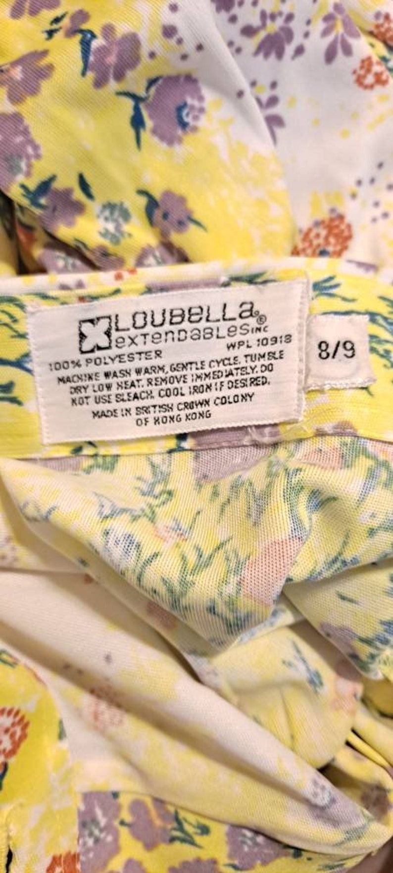 May include: A yellow and white floral fabric with a white tag that reads "Loubella extendables inc. WPL 10918 100% polyester Machine wash warm, gentle cycle. Tumble dry low heat. Remove immediately. Do not use bleach. Cool iron if desired. Made in British Crown Colony of Hong Kong 8/9"