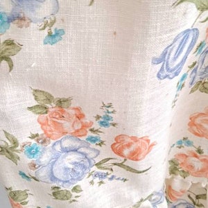 Puede incluir: Una tela blanca con un patr&oacute;n floral con flores azules, rosas y verdes. La tela tiene un borde con volantes.