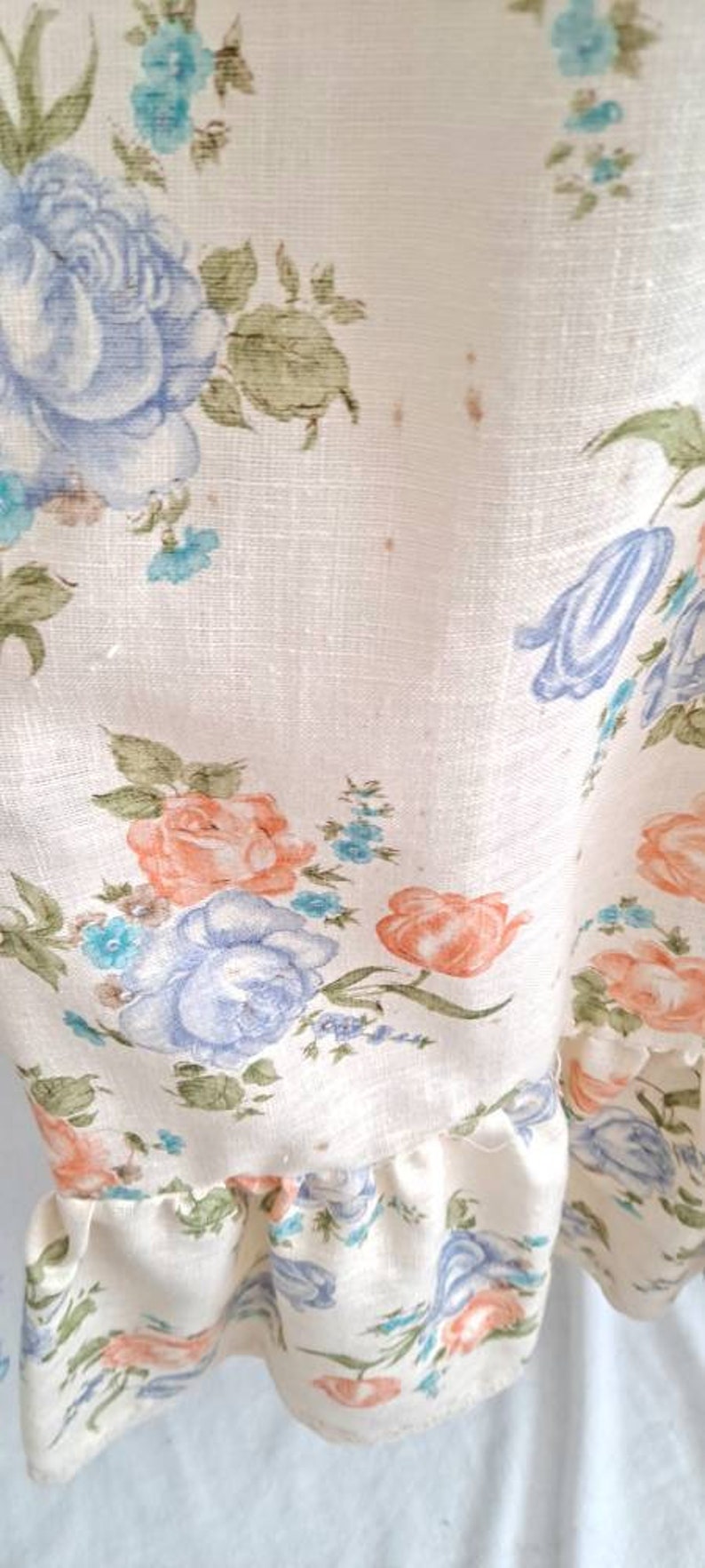 Puede incluir: Una tela blanca con un patr&oacute;n floral con flores azules, rosas y verdes. La tela tiene un borde con volantes.