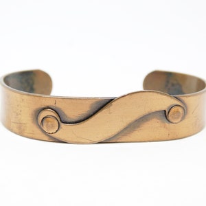 May include: A copper bracelet with a wavy design and two round accents. The bracelet is made of hammered metal and has a rustic look.