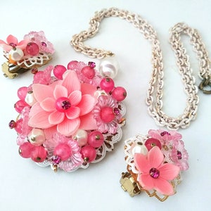 May include: A pink flower necklace and earrings set with white pearl accents. The necklace has a delicate chain and the earrings are clip-on.