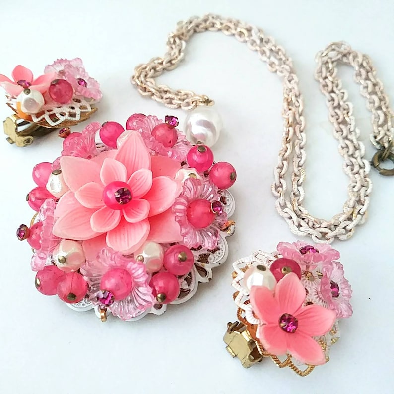 May include: A pink flower necklace and earrings set with white pearl accents. The necklace has a delicate chain and the earrings are clip-on.