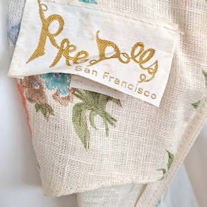 Puede incluir: Una tela blanca con estampado floral y una etiqueta dorada que dice "Ree Dolls San Francisco".