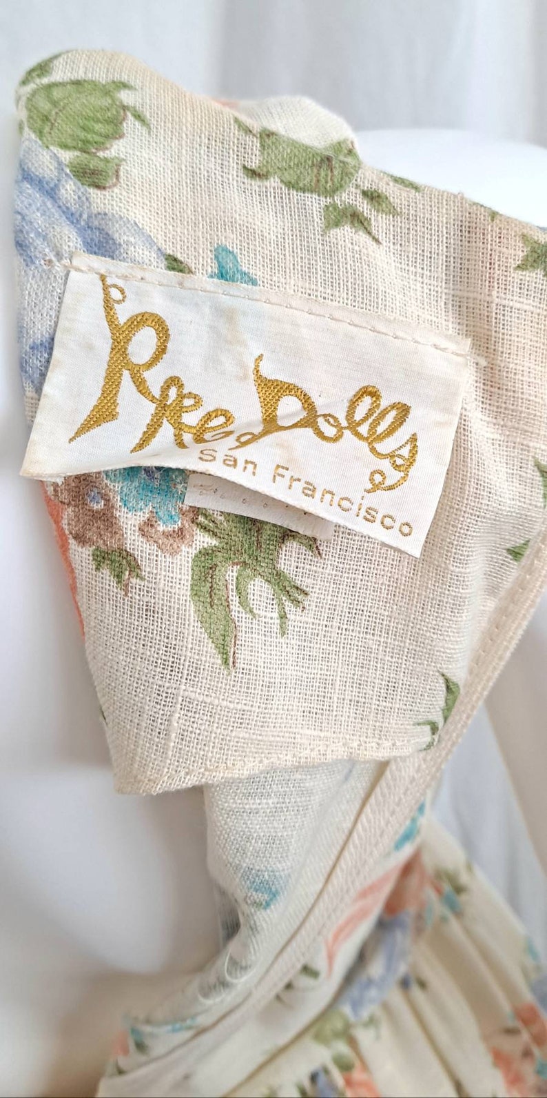 Puede incluir: Una tela blanca con estampado floral y una etiqueta dorada que dice "Ree Dolls San Francisco".