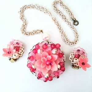 May include: A pink and white flower-shaped necklace and earrings set. The necklace has a large pendant with a pink flower surrounded by pink and white beads. The earrings are smaller versions of the pendant.