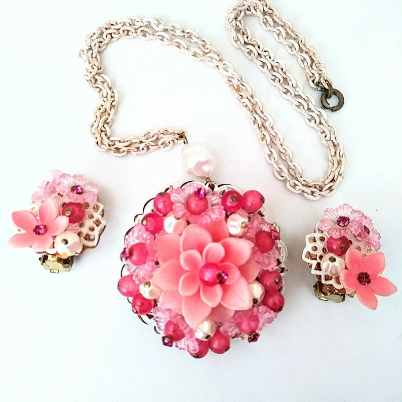 May include: A pink and white flower-shaped necklace and earrings set. The necklace has a large pendant with a pink flower surrounded by pink and white beads. The earrings are smaller versions of the pendant.
