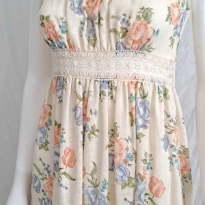 Puede incluir: Un vestido maxi sin mangas con estampado floral en tonos azules, rosas y verdes. El vestido tiene un ribete de encaje blanco alrededor del escote y la cintura.