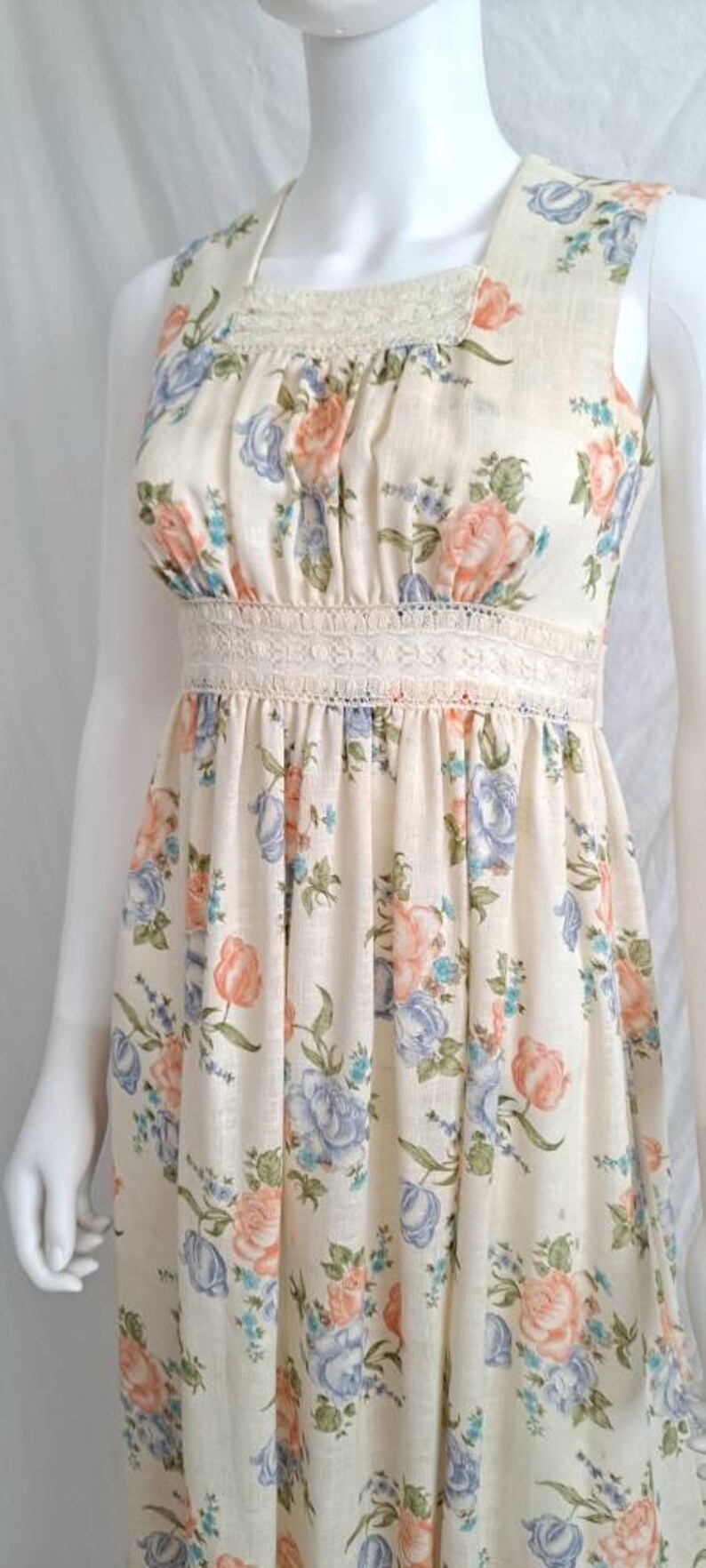 Puede incluir: Un vestido maxi sin mangas con estampado floral en tonos azules, rosas y verdes. El vestido tiene un ribete de encaje blanco alrededor del escote y la cintura.