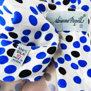 May include: White silk blouse with blue and black polka dots. The blouse has a label that reads "Adrianna Papell" and a tag that says "100% Silk, 10, Made in Korea."