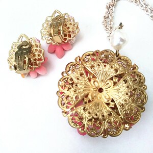 May include: A gold tone necklace and earrings set with pink flower accents. The necklace features a large pendant with a filigree design and a pearl drop.