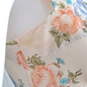 Puede incluir: Un vestido de lino blanco con estampado floral con flores rosas, azules y verdes. El vestido tiene un ribete de encaje y un dobladillo con volantes.
