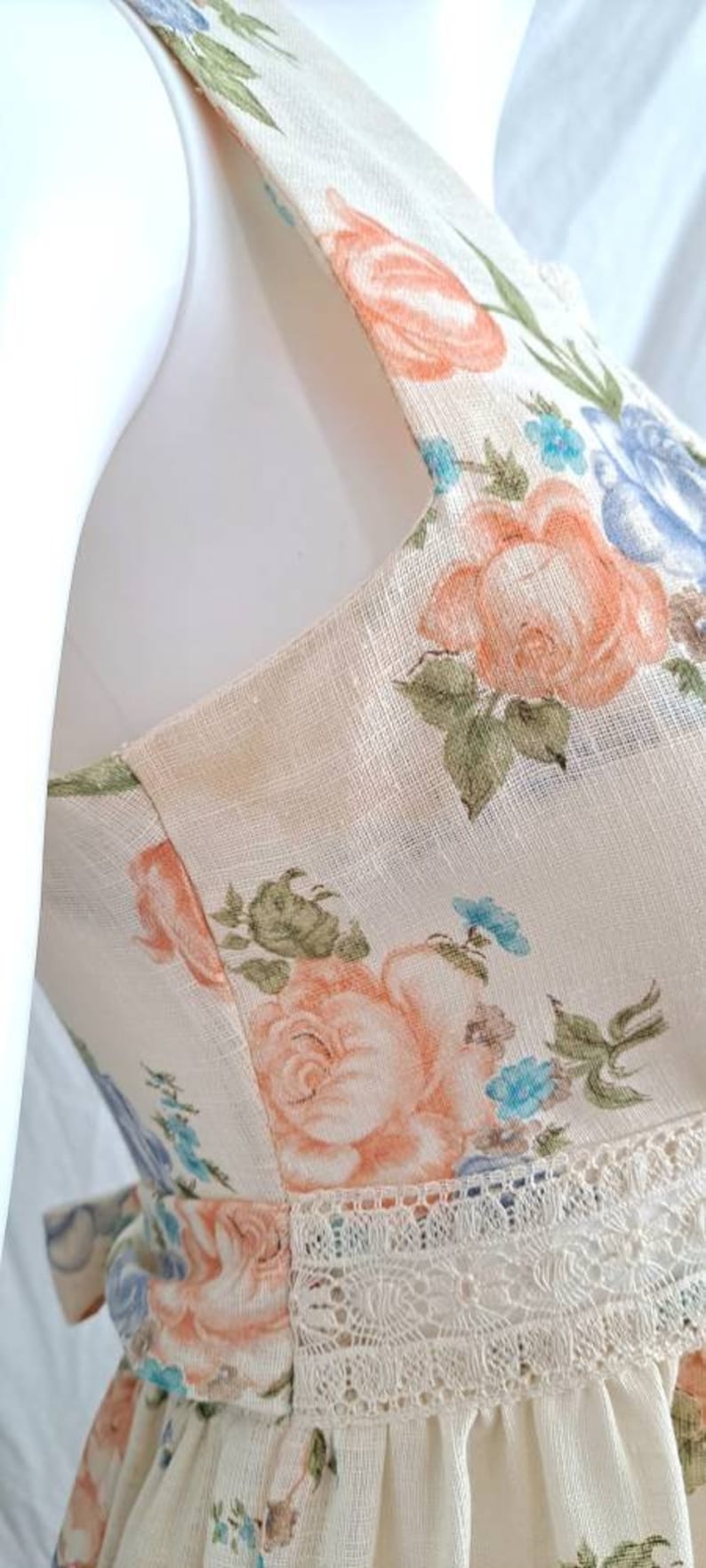 Puede incluir: Un vestido de lino blanco con estampado floral con flores rosas, azules y verdes. El vestido tiene un ribete de encaje y un dobladillo con volantes.