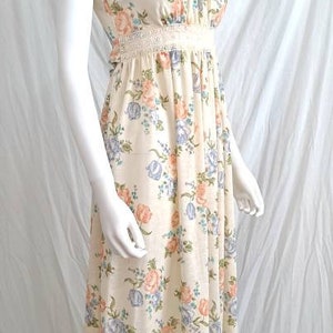 Puede incluir: Un vestido largo blanco maxi con estampado floral con flores rosas, azules y amarillas. El vestido tiene un ribete de encaje en el escote y la cintura. El vestido tiene un dobladillo con volantes.