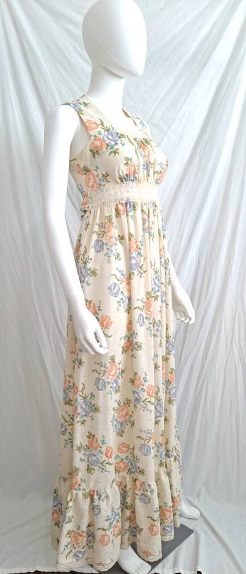 Puede incluir: Un vestido largo blanco maxi con estampado floral con flores rosas, azules y amarillas. El vestido tiene un ribete de encaje en el escote y la cintura. El vestido tiene un dobladillo con volantes.