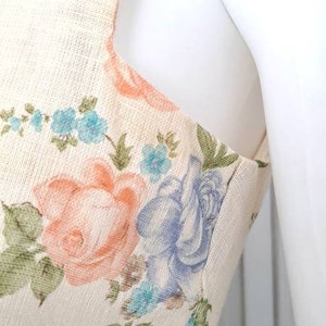 Puede incluir: Un vestido sin mangas con estampado floral en tonos azules, rosas y verdes. El vestido tiene un ribete de encaje en la parte inferior y un fondo crema.