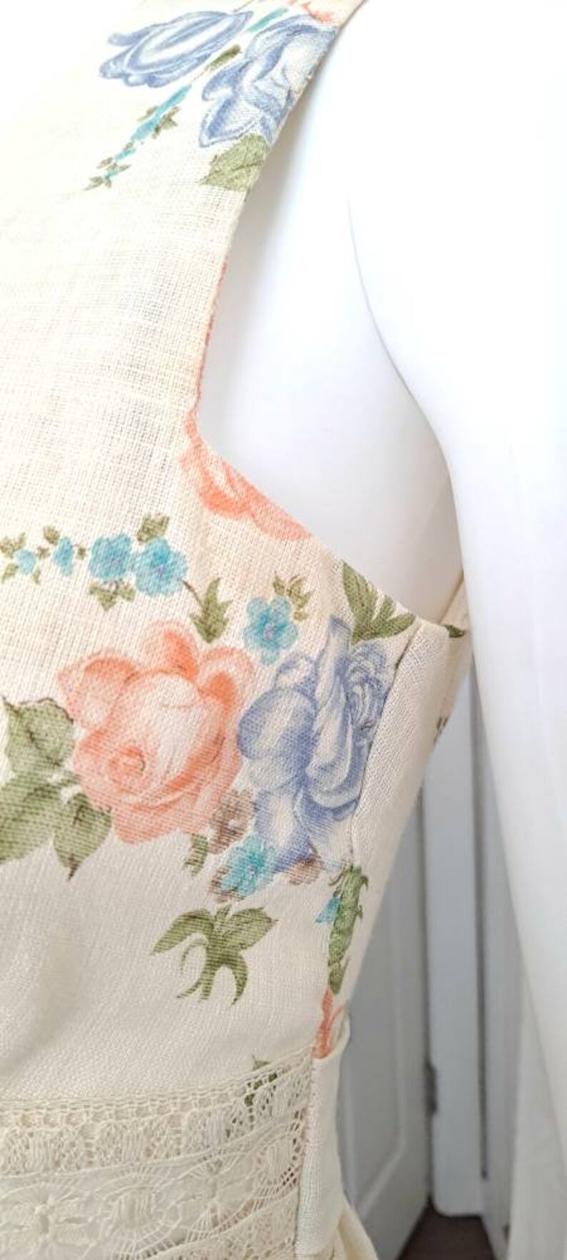 Puede incluir: Un vestido sin mangas con estampado floral en tonos azules, rosas y verdes. El vestido tiene un ribete de encaje en la parte inferior y un fondo crema.