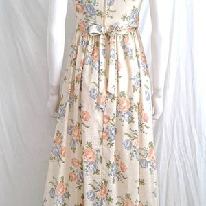 Puede incluir: Un vestido largo blanco con estampado floral con flores rosas, azules y verdes. El vestido tiene un corpi&ntilde;o ajustado y una falda fluida con un dobladillo con volantes.