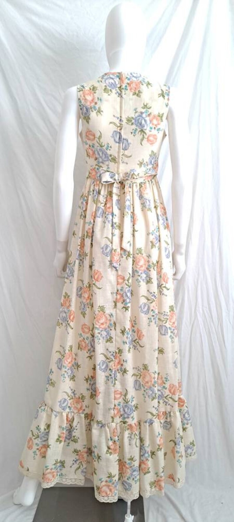 Puede incluir: Un vestido largo blanco con estampado floral con flores rosas, azules y verdes. El vestido tiene un corpi&ntilde;o ajustado y una falda fluida con un dobladillo con volantes.