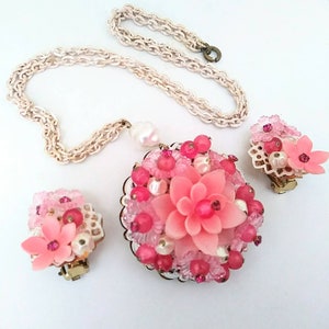 May include: A pink and white flower-shaped necklace and earrings set. The necklace has a delicate chain and a large pendant with a pink flower surrounded by pink and white beads. The earrings are clip-on and feature a smaller pink flower with pink and white beads.