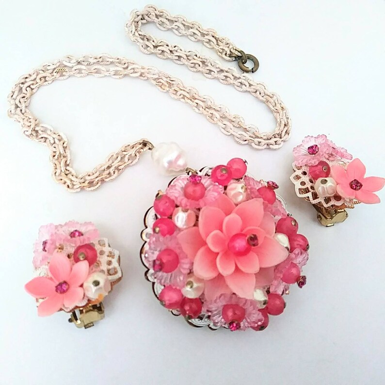 May include: A pink and white flower-shaped necklace and earrings set. The necklace has a delicate chain and a large pendant with a pink flower surrounded by pink and white beads. The earrings are clip-on and feature a smaller pink flower with pink and white beads.
