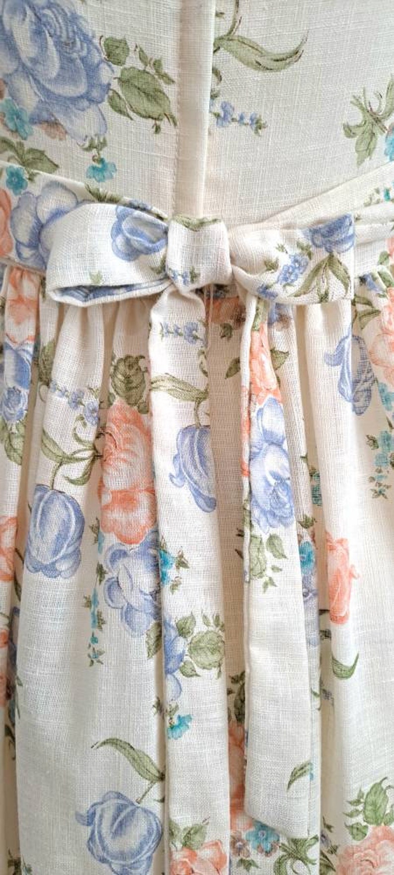 Puede incluir: Un vestido blanco con estampado floral con flores azules y rosas. El vestido tiene un lazo en la cintura.
