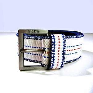 Könnte beinhalten: Ein weißer Canvas-Gürtel mit blauen und roten Streifen und einer silbernen Schnalle. Die Schnalle trägt die Gravur "OGBETTI".