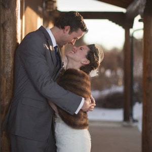 May include: A bride and groom embrace in a winter setting. The bride is wearing a white wedding dress with a brown fur shawl and a veil. The groom is wearing a grey suit and a white shirt. They are standing in front of a wooden structure.