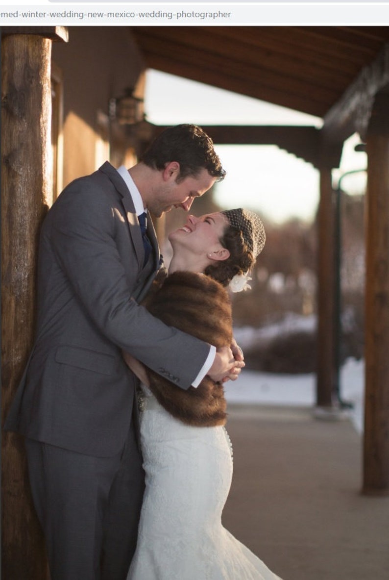 May include: A bride and groom embrace in a winter setting. The bride is wearing a white wedding dress with a brown fur shawl and a veil. The groom is wearing a grey suit and a white shirt. They are standing in front of a wooden structure.