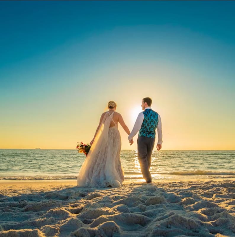 May include: A bride and groom in wedding attire walk hand-in-hand on a sandy beach towards the ocean. The sun is setting in the background, casting a golden glow over the scene. The bride is wearing a white wedding dress with a long veil, and the groom is wearing a blue vest and gray pants.