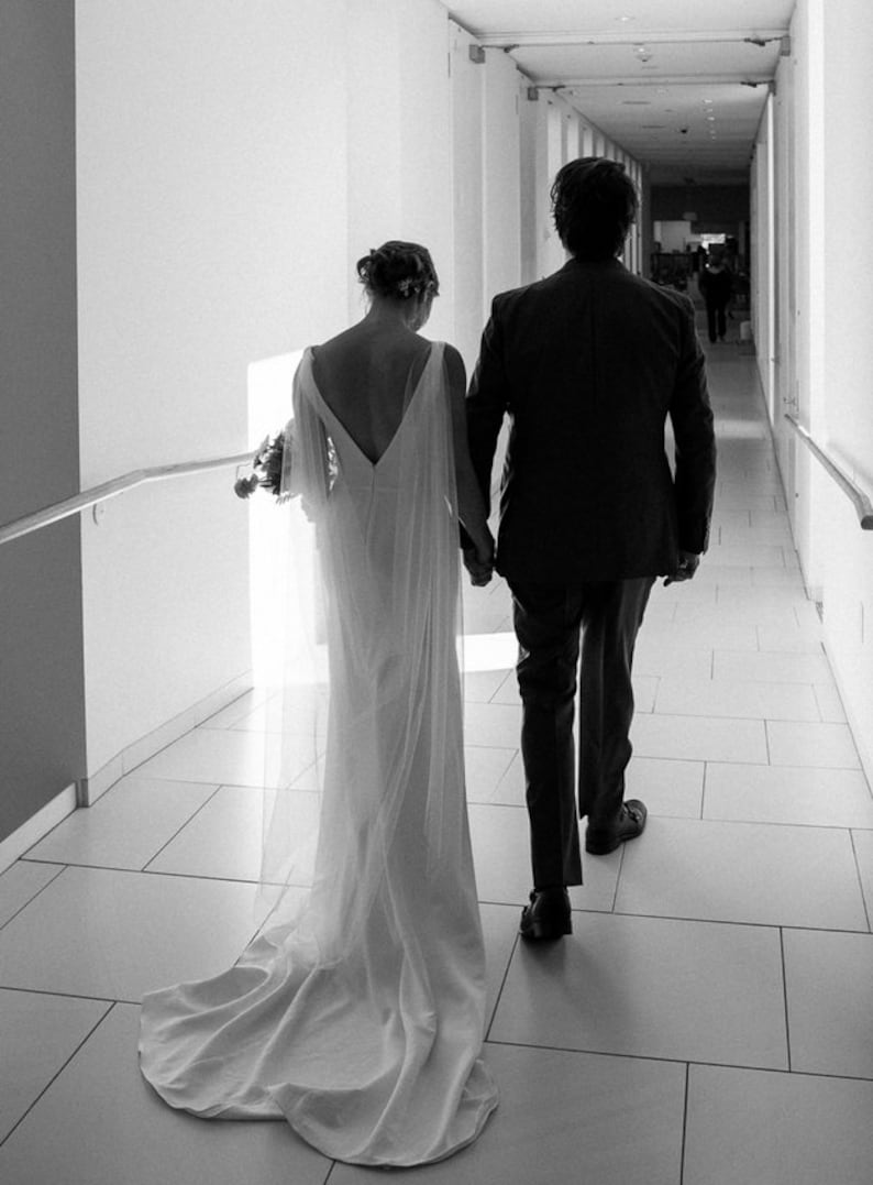 May include: A bride and groom in a white wedding dress and a black suit walk down a white tiled hallway.