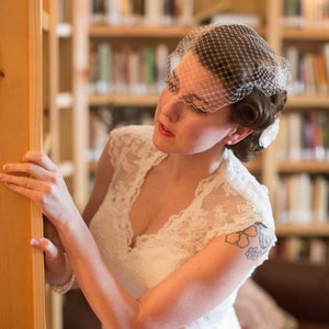 May include: A bride wearing a white lace wedding dress with a veil. She is standing in front of a bookshelf with a wooden door.