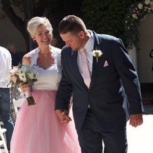 May include: A bride and groom walking down the aisle. The bride is wearing a white lace top with a pink tulle skirt. The groom is wearing a dark blue suit with a pink tie.