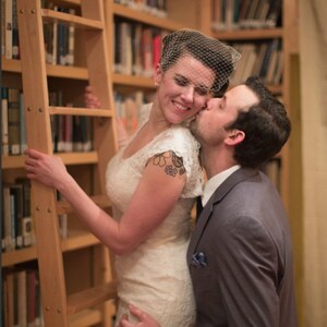 May include: A couple pose for a wedding photograph in a library. The bride is wearing a white lace dress and a veil. The groom is wearing a grey suit and a blue tie. They are standing in front of a bookcase filled with books.