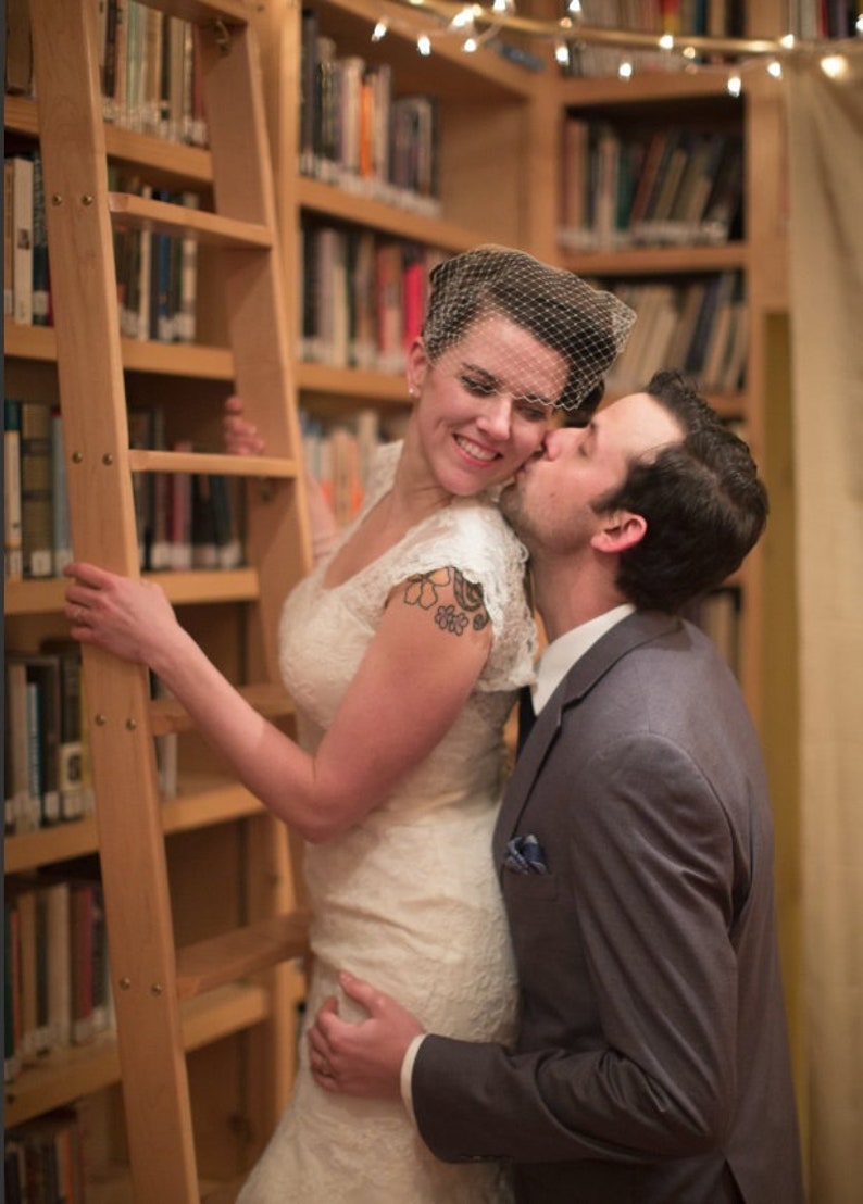 May include: A couple pose for a wedding photograph in a library. The bride is wearing a white lace dress and a veil. The groom is wearing a grey suit and a blue tie. They are standing in front of a bookcase filled with books.